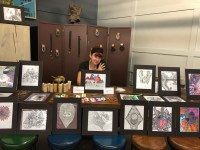 Artists Lisa Cameron sits behind her art display. Her art include multiple framed ink drawings, jewelery and homemade lotion